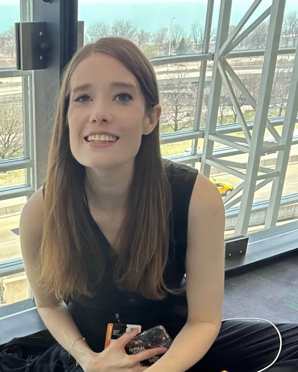Vicki smiling beside tall convention-center windows with a conference badge in hand.