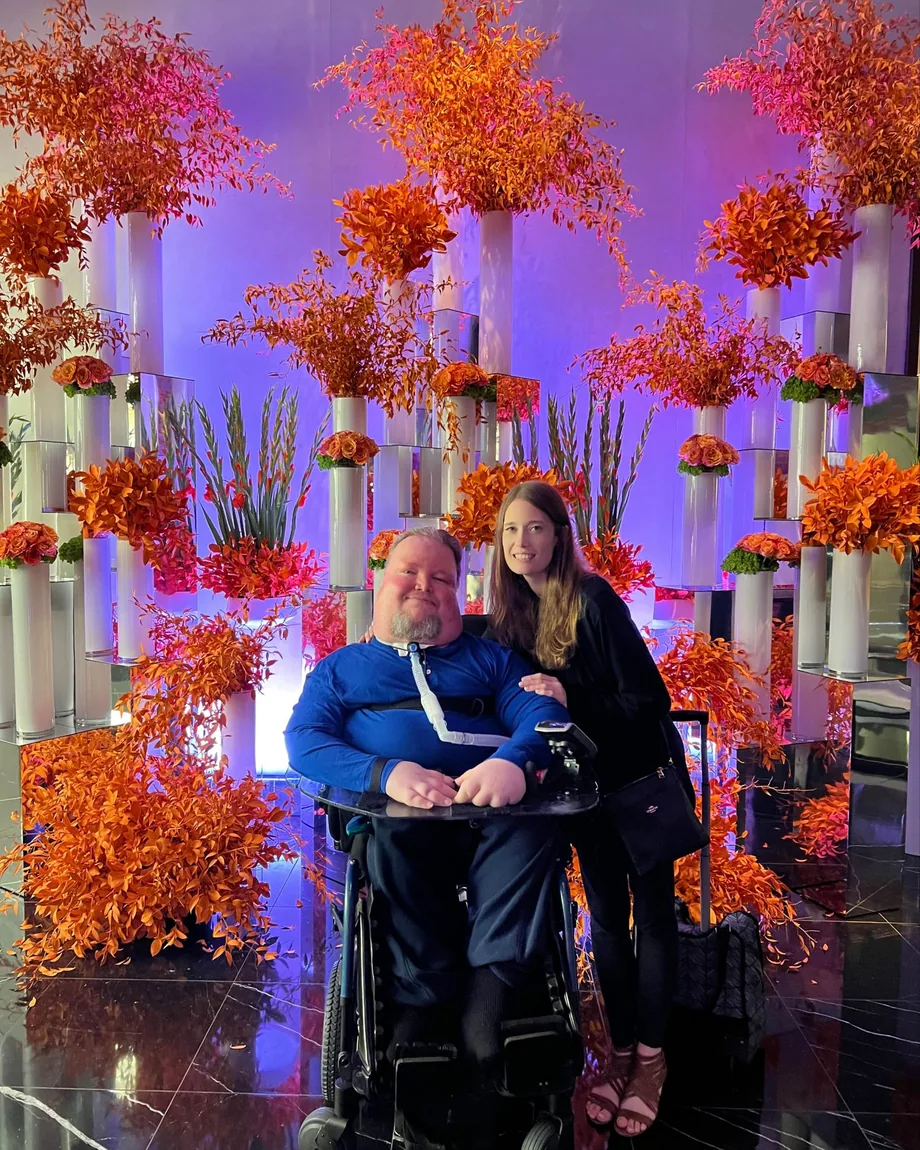 Vicki and Steve smiling together in front of an orange flower wall under purple light.