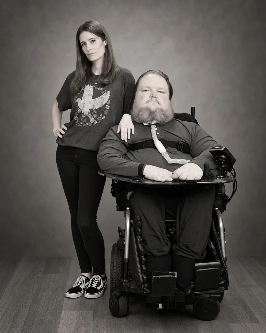 Black-and-white studio portrait of Vicki standing beside Steve in his wheelchair, her hand on his shoulder.